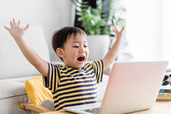 Kind sitzt vor dem Laptop und hebt die Arme in die Höhe.